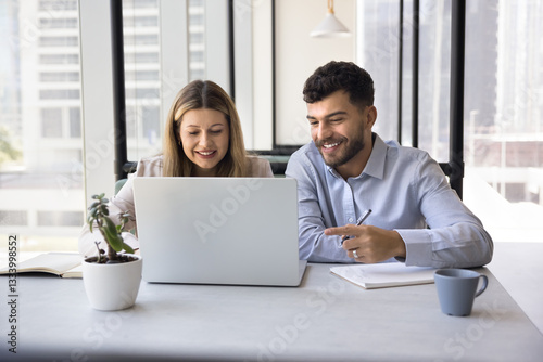 In open-space office two teammates working on joint online project, review new program on laptop, smile while watching content on internet, prepare presentation feel satisfied with productive workflow