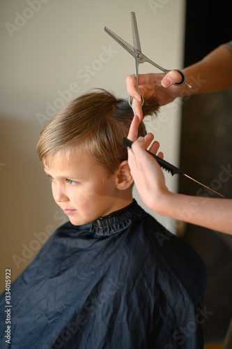 A little blond boy gets a haircut, sitting in a salon chair with a cape. The hairdresser holds scissors and a comb. The child is calm and enjoying the experience. Concept of childhood grooming and sel