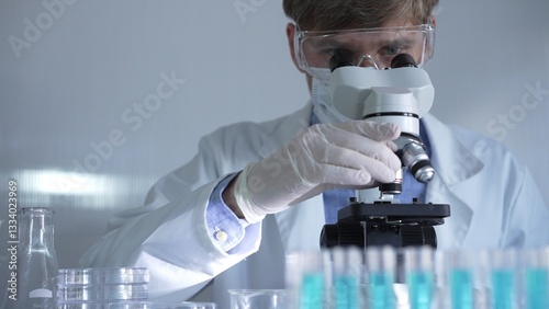 Male scientist, wearing a lab coat, mask, white gloves and safety glasses, is using a binocular microscope surrounded by test tubes in laboratory