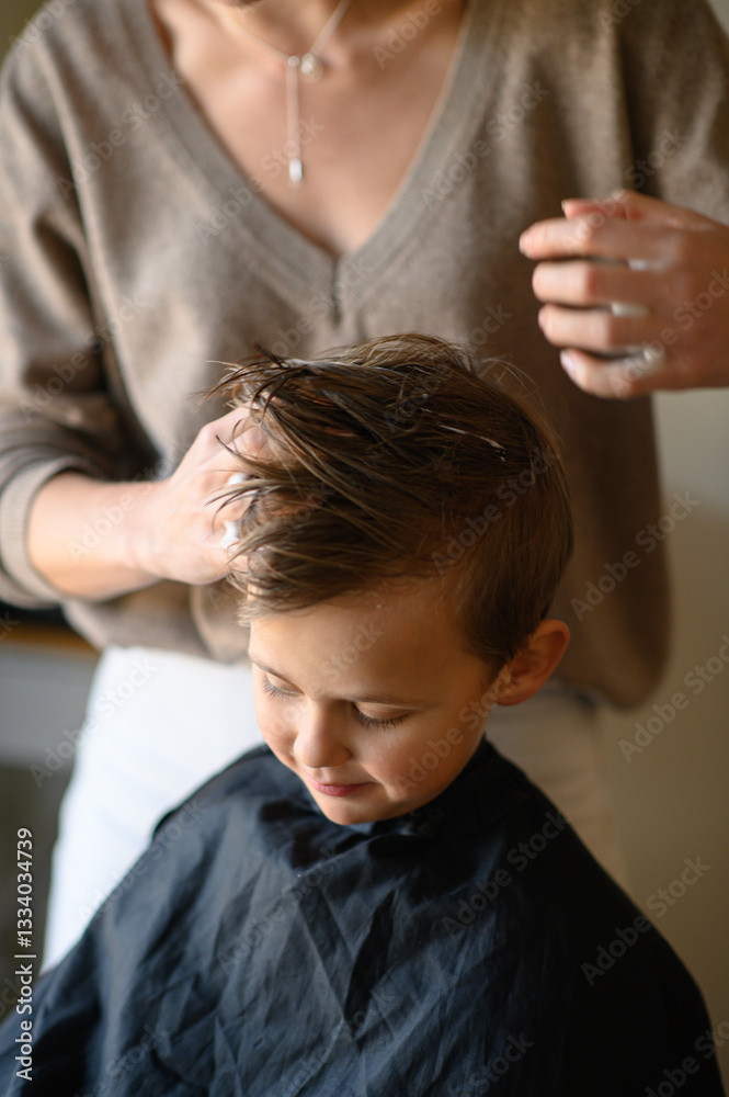 Fototapeta premium A little blond boy enjoys his new haircut as the hairdresser styles his hair with mousse. He is happy, laughing, and having fun in the salon chair. Concept of childhood grooming and self-care.