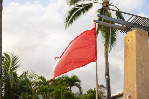 A tattered red warning flag waving in the wind, set against palm trees and a partly cloudy sky. Cautionary symbol for danger, high winds, or hazardous conditions