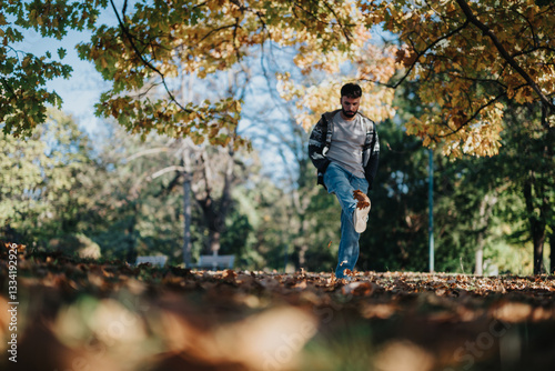 A young man wearing a casual jacket and jeans playfully kicks autumn leaves in a sunlit park. Vibrant foliage surrounds him, creating a serene and joyful atmosphere perfect for relaxation.