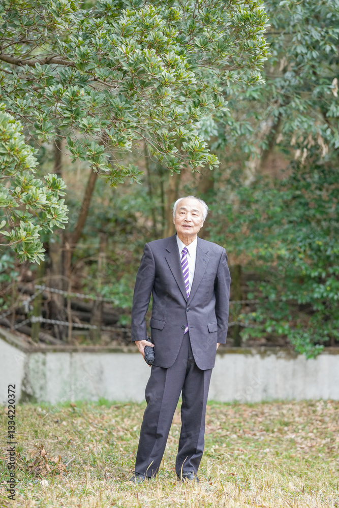 A park on a pleasant spring daytime with plenty of greenery in Japan. A Japanese man in his late 70s wearing a suit stands up. He has a folding umbrella. A park on a pleasant spring daytime with plenty of greenery in Japan. A Japanese man in his late 70s wearing a suit stands up. He has a folding umbrella.