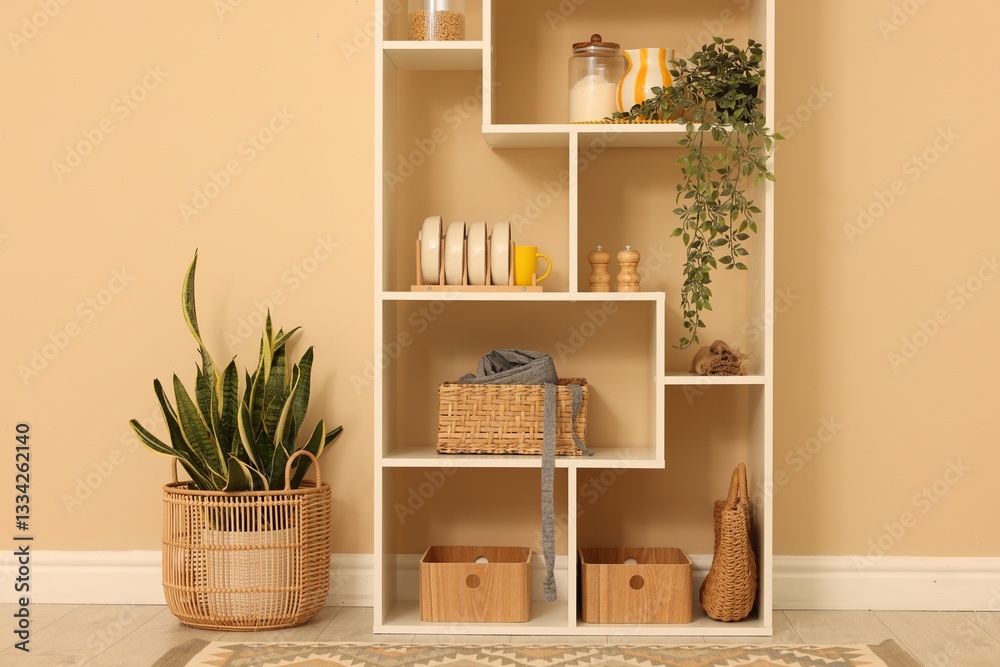 Fototapeta premium Shelving unit with dishware, jars of different products and other stuff near beige wall indoors