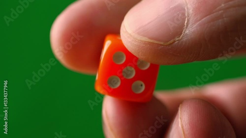 A close-up shot of a hand holding an orange die against a green background. The focus is on the die and the fingers holding it, showcasing a moment of chance or decision