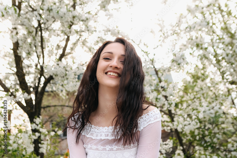 Cheerful young woman enjoying sunny spring day in blooming garden, posing against blossoming trees, wearing white dress, looking happy and relaxed Cheerful young woman enjoying sunny spring day in blooming garden, posing against blossoming trees, wearing white dress, looking happy and relaxed