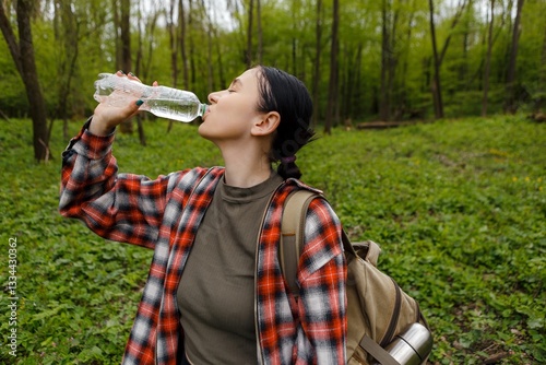 Female hiker drinking water from bottle, resting amid lush forest greenery during solitary wilderness trek, feeling energized by nature's tranquil surroundings