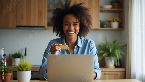 Happy excited young African woman customer winner holding credit card using laptop computer in kitchen celebrating money win receiving cashback getting online bonus for shopping sitting at home. conce