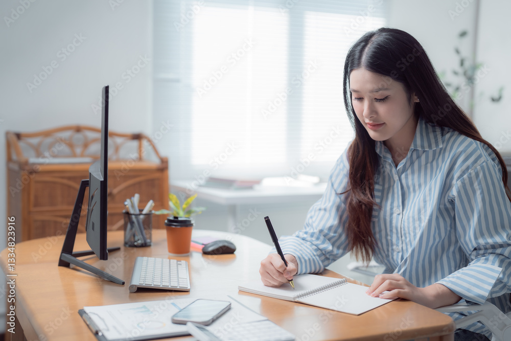 Young Asian businesswoman writing notes in empty notebook while sitting at her desk in modern workplace, surrounded by computer, keyboard, mouse, smartphone, calculator and documents Young Asian businesswoman writing notes in empty notebook while sitting at her desk in modern workplace, surrounded by computer, keyboard, mouse, smartphone, calculator and documents