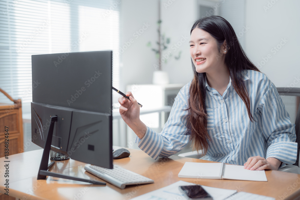 Young asian businesswoman pointing with a pen at computer monitor while working from home office and taking notes on a notepad during a video call meeting Young asian businesswoman pointing with a pen at computer monitor while working from home office and taking notes on a notepad during a video call meeting