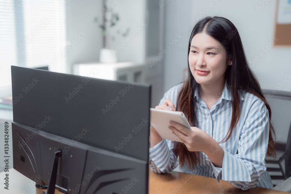 Young Asian businesswoman smiling while taking notes on a notepad, focused on the computer screen in her bright home office, creating a productive and comfortable work environment Young Asian businesswoman smiling while taking notes on a notepad, focused on the computer screen in her bright home office, creating a productive and comfortable work environment