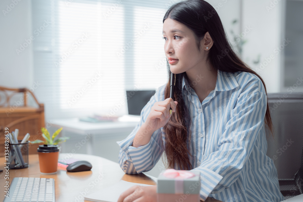 Young asian businesswoman is thinking about something while working with computer at modern office with gift box on desk, celebrating birthday or special occasion at workplace Young asian businesswoman is thinking about something while working with computer at modern office with gift box on desk, celebrating birthday or special occasion at workplace