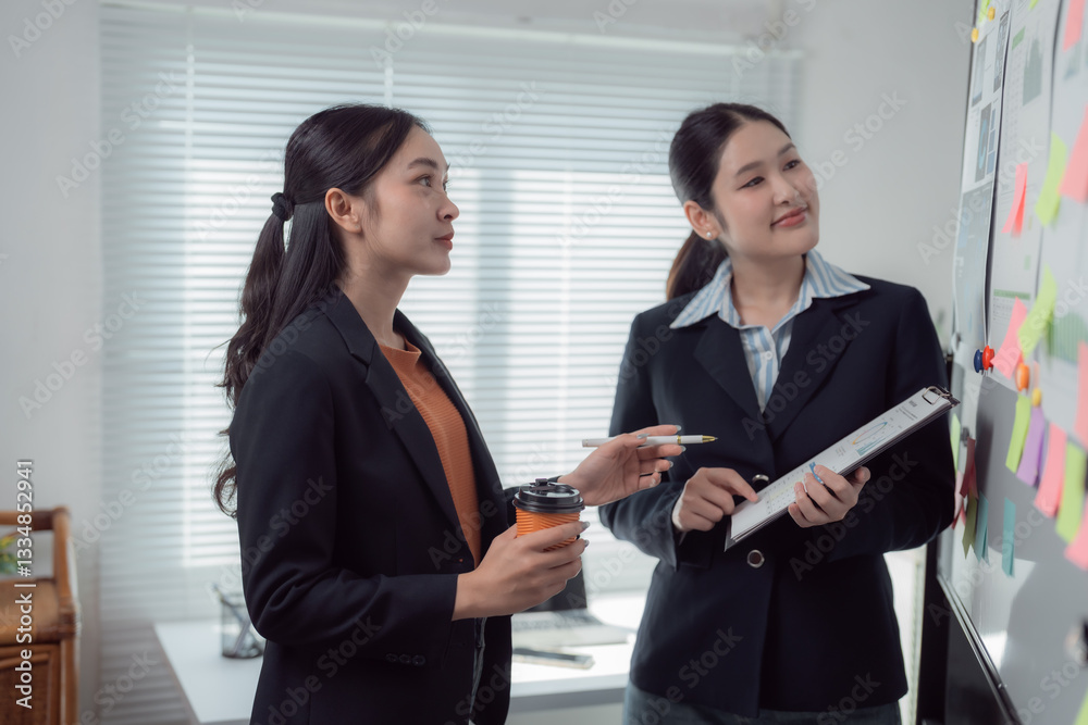 Two asian businesswomen are discussing company strategy with sticky notes attached to a whiteboard in a modern office, holding a takeaway coffee and a clipboard Two asian businesswomen are discussing company strategy with sticky notes attached to a whiteboard in a modern office, holding a takeaway coffee and a clipboard