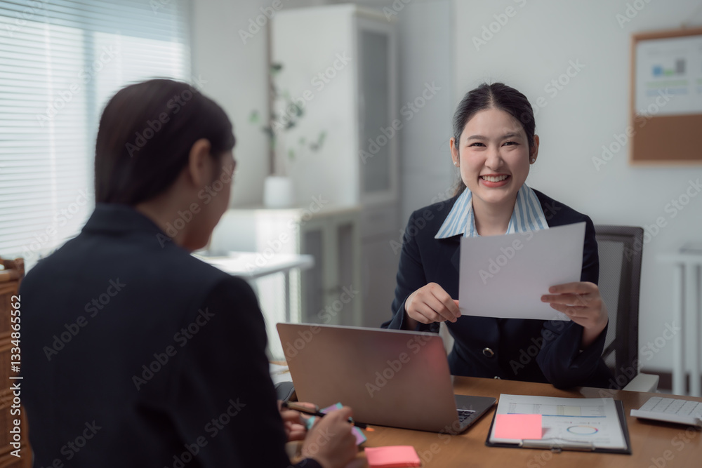 Two businesswomen are having a meeting in a modern office, one of them is smiling and showing a document to her colleague who's taking notes Two businesswomen are having a meeting in a modern office, one of them is smiling and showing a document to her colleague who's taking notes