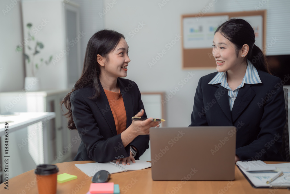 Two asian businesswomen are discussing marketing strategy using laptop and taking notes in modern office, collaborating on a project and sharing ideas Two asian businesswomen are discussing marketing strategy using laptop and taking notes in modern office, collaborating on a project and sharing ideas