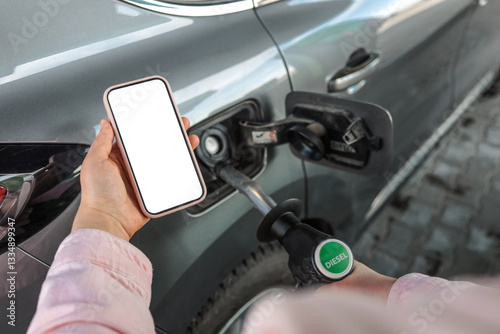 Person refueling a car with diesel pump while holding a smartphone with blank screen. Concept of refueling, transport, diesel fuel, gas station, mobile app, and payment