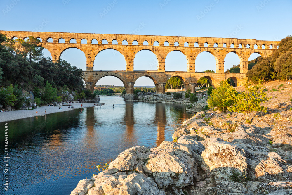 Fototapeta premium Pont du Gard, ancient Roman aqueduct bridge, Provence late afternoon golden light on arches mirroring in river, Gorges du Gardon, natural landscape designated Biosphere Reserve by UNESCO, France