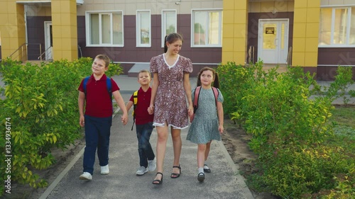 Mother leading children from school, female mother holding children hand, happy family, young nanny with children, schoolchildren with backpacks walking with aunt by the hand, little boys girl walking
