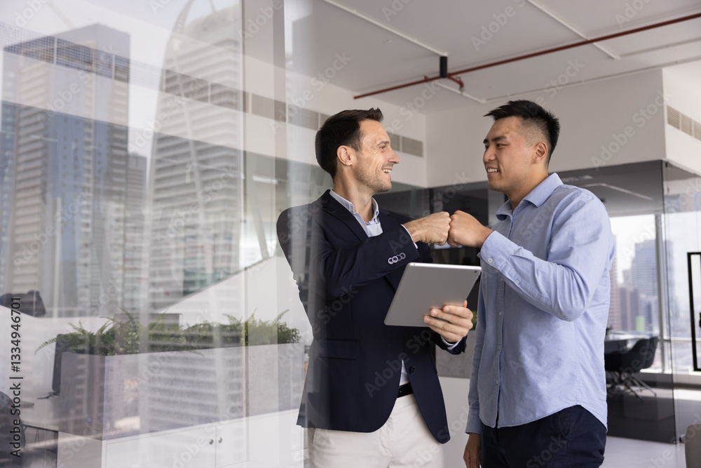 Fototapeta premium Two positive male business colleagues enjoying partnership, friendship, giving fist bump due to successful effective teamwork, using tablet computer, standing in office, talking, laughing