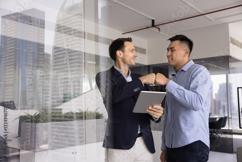 Two positive male business colleagues enjoying partnership, friendship, giving fist bump due to successful effective teamwork, using tablet computer, standing in office, talking, laughing
