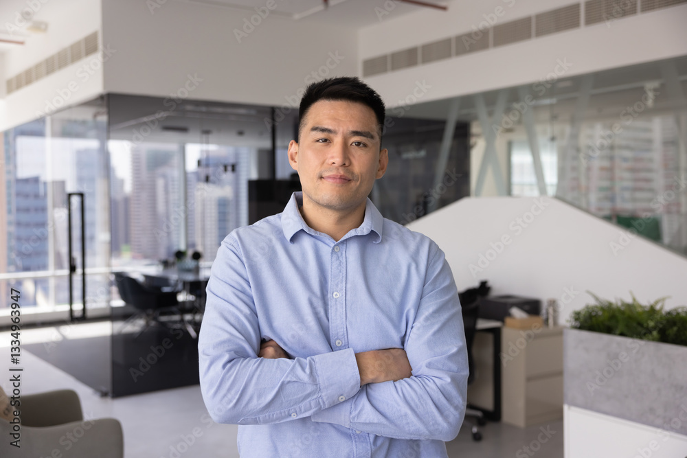 Fototapeta premium Confident successful young Asian company CEO man posing for business portrait, looking at camera with hands folded in confidence gesture, standing in modern office workspace alone