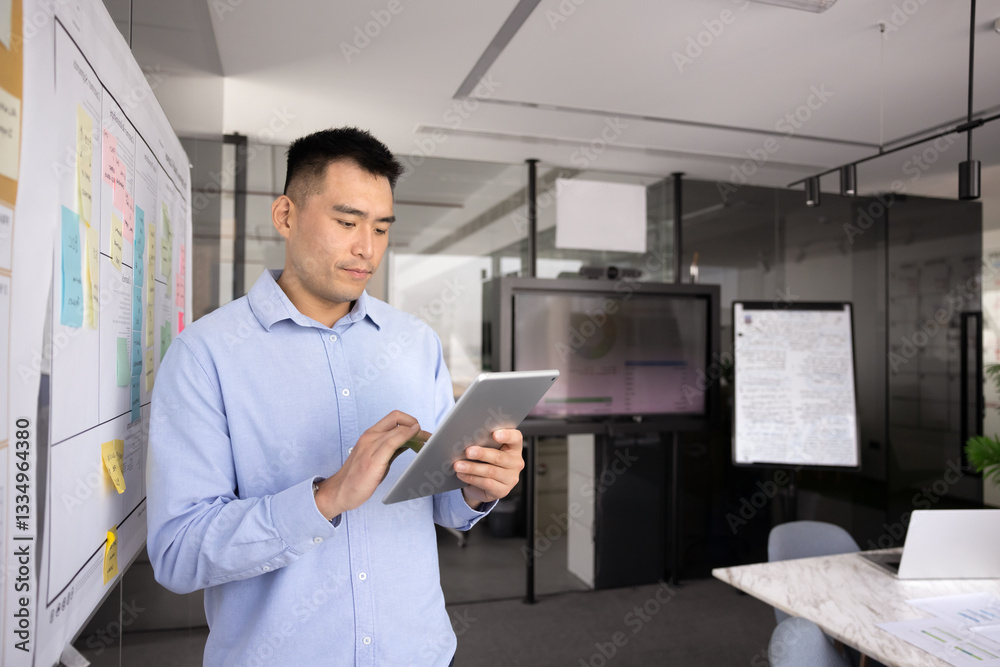 Focused young Asian manager man using modern technology on tablet computer in office meeting workspace alone, standing at whiteboard with stickers, working on online application Focused young Asian manager man using modern technology on tablet computer in office meeting workspace alone, standing at whiteboard with stickers, working on online application