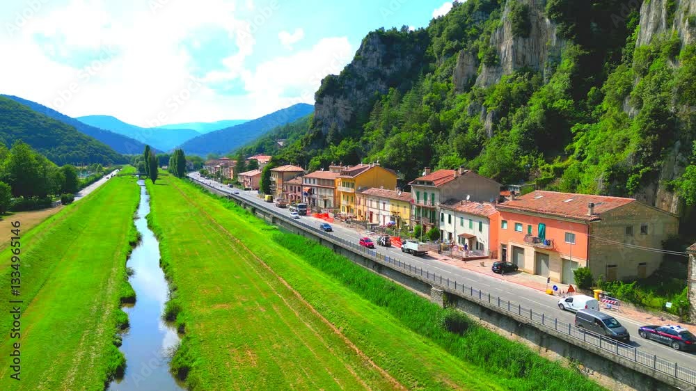 Gorgeous aerial pedestal down shot from Pioraco, a gem valley nested inside the rocky green Sibillini mountains, featuring the mirroring Potenza river, meadows, trees, road with cars, some buildings
