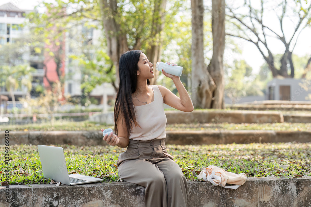 Naklejka premium Sustainable lifestyle. Young woman enjoying a refreshing drink while working on her laptop outdoors.