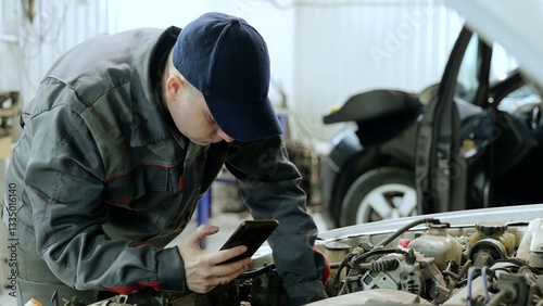 Experienced automotive technician checking smartphone while diagnosing car engine problems in modern repair workshop, focusing on precise mechanical diagnostics photo