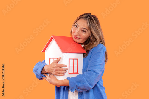 Smiling woman holds a miniature house model against a vibrant orange background during a cheerful moment expressing love for home photo