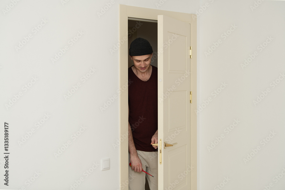 A man wearing a beanie and casual clothes stands at the door frame, holding tools, as he prepares to enter a bright, minimalistic room. The decor is simple, allowing natural light A man wearing a beanie and casual clothes stands at the door frame, holding tools, as he prepares to enter a bright, minimalistic room. The decor is simple, allowing natural light
