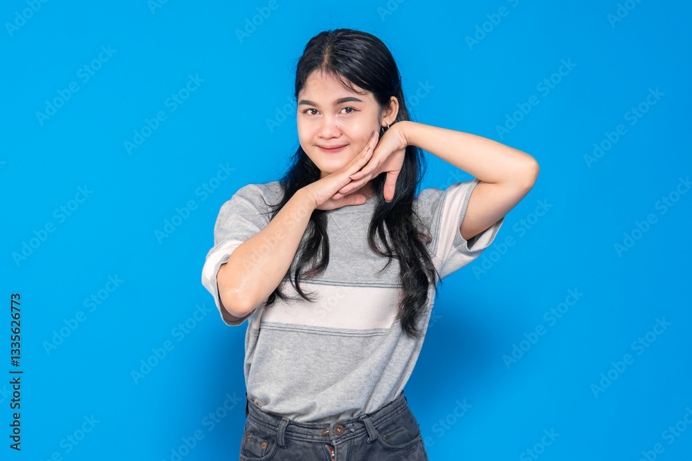 Young Asian woman posing playfully with a charming smile, resting her face on her hands. She exudes happiness and confidence against a vibrant blue background. Young Asian woman posing playfully with a charming smile, resting her face on her hands. She exudes happiness and confidence against a vibrant blue background.