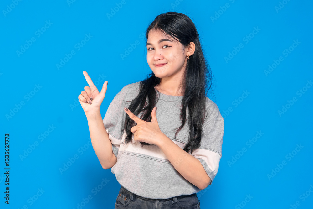 Asian young woman with long black hair wearing a gray shirt, smiling playfully while pointing with both hands in a finger-gun gesture against a blue background. Asian young woman with long black hair wearing a gray shirt, smiling playfully while pointing with both hands in a finger-gun gesture against a blue background.
