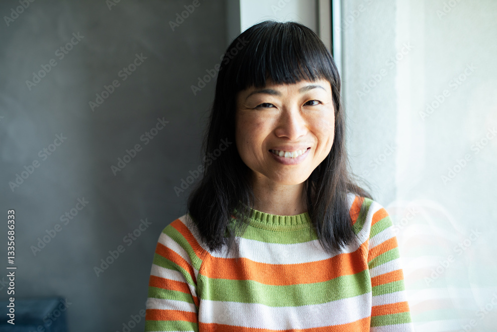 Smiling portrait of a Asian woman wearing striped sweater standing by a window Smiling portrait of a Asian woman wearing striped sweater standing by a window