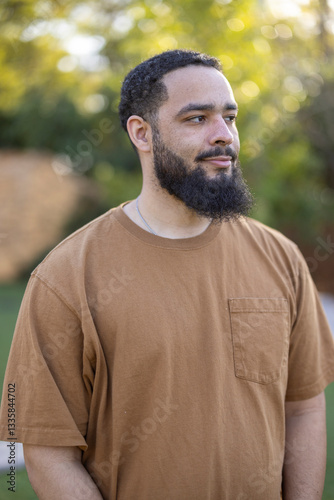 A confident man with a stylish beard wears a soft brown tshirt, exuding a relaxed vibe amid lush greenery and warm sunlight, embodying a cheerful and positive demeanor that makes him stand out