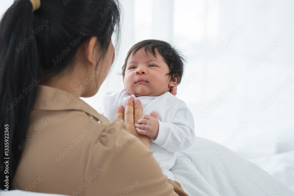Portrait of multiracial newborn baby lying in arm of Asian mother who touching her chest with gentle. Little cute infant looking at mom with innocent eyes on bed at home Portrait of multiracial newborn baby lying in arm of Asian mother who touching her chest with gentle. Little cute infant looking at mom with innocent eyes on bed at home