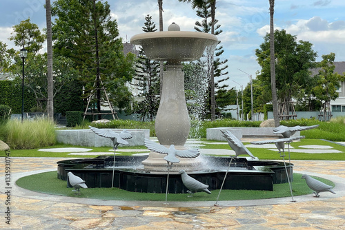 Outdoor image of a fountain sculpture in a small public park show water drops by a summer breeze with trees and shrubs in countryside suburb area.
