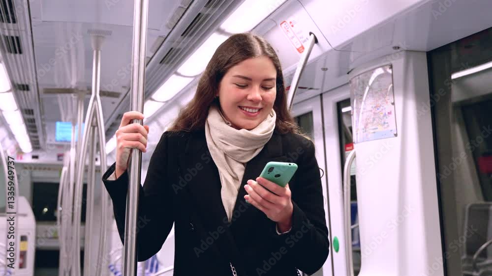 Young Caucasian woman holding a smartphone and standing in a subway train, smiling softly while gripping a pole, wearing a black coat and scarf during an urban commute in public transportation.