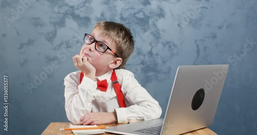 Young pupil joyfully daydreams in front of laptop at desk. Cheerful schoolboy in glasses ponders deeply with notepad and laptop on table