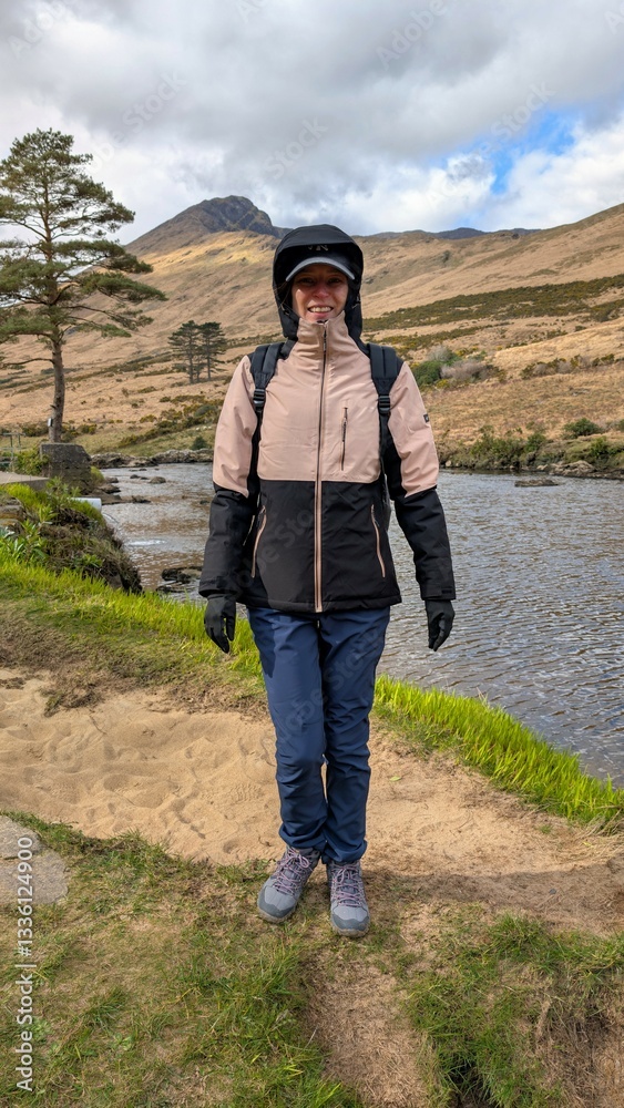 Naklejka premium Beautiful woman backpacker in hooded jacket hiking at river Erriff near Aesleagh, county Mayo, Ireland, Irish landscape, mountains and nature background 