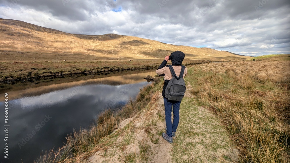 Beautiful woman backpacker in hooded jacket hiking at river Erriff near Aesleagh, county Mayo, Ireland, Irish landscape, mountains and nature background