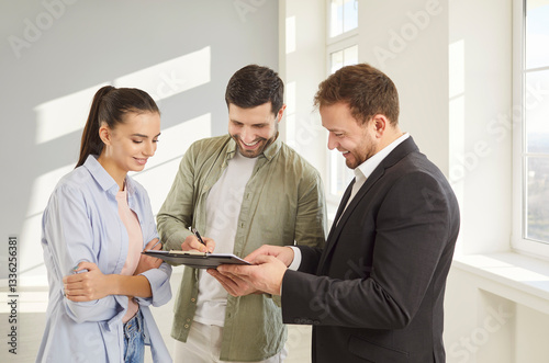 Family couple buyers signing contract document in real estate agent after buying new apartment, put signature on papers, standing in empty room near window. Loan agreement or mortgage