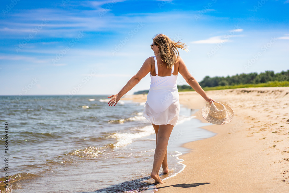 Beautiful middle-aged woman in swimsuit holding hat walking on sandy beach in summertime. Energy and happiness. Back view Beautiful middle-aged woman in swimsuit holding hat walking on sandy beach in summertime. Energy and happiness. Back view