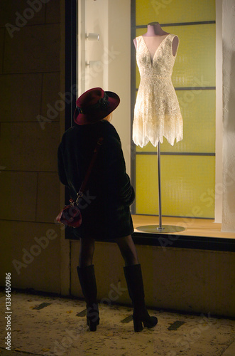 Elegant woman with red hat and red handbag stands in front of an illuminated shop window at night, in which a mannequin wearing a sexy white short dress with a plunging neckline is standing