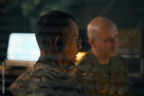 Two military officers engaged in monitoring operations within command center environment. One officer wears headset and looks focused on task at hand