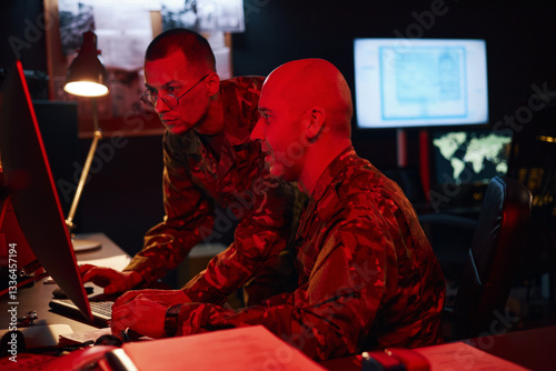 Two military personnel examining computer screens in command center with multiple monitors displaying various data. One person is viewing a document while the other looks at the screen