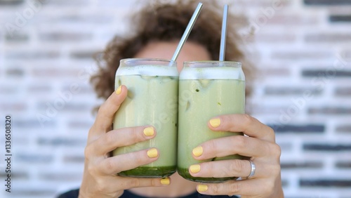 Caucasian woman with curly hair and yellow nail polish holding two glasses of iced matcha latte with metal straws against a brick wall background
