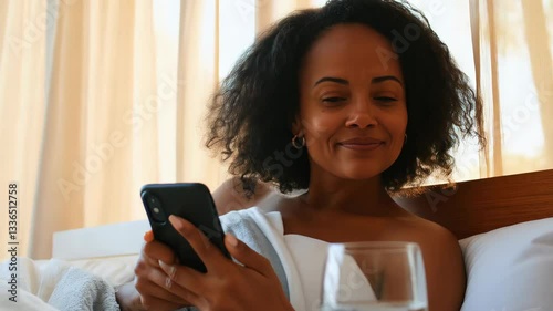 Smiling woman relaxing in bed with a smartphone during a peaceful morning at home
