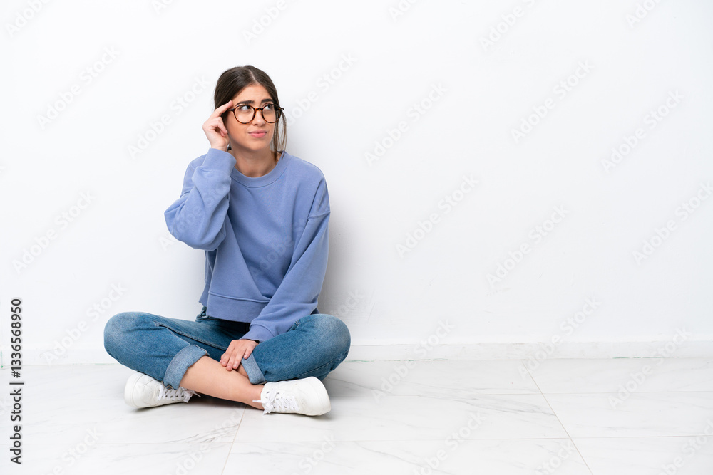 Young caucasian woman sitting on the floor isolated on white background having doubts and with confuse face expression Young caucasian woman sitting on the floor isolated on white background having doubts and with confuse face expression