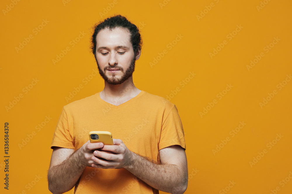 Young man using a smartphone against a vibrant yellow background, wearing a casual orange t shirt, showcasing a focused expression, ideal for technology and communication themes Young man using a smartphone against a vibrant yellow background, wearing a casual orange t shirt, showcasing a focused expression, ideal for technology and communication themes